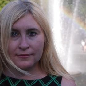 Photo of Kristen Gillespie-Lynch, woman with blonde hair and diagonal striped top in front of rainbow and water background