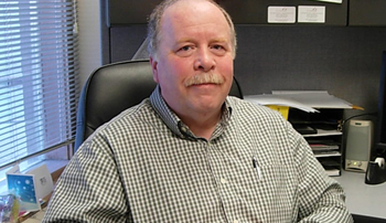 Photo of Mark A. Smith, a white man with a mustache. He is smiling at the camera from behind his desk.