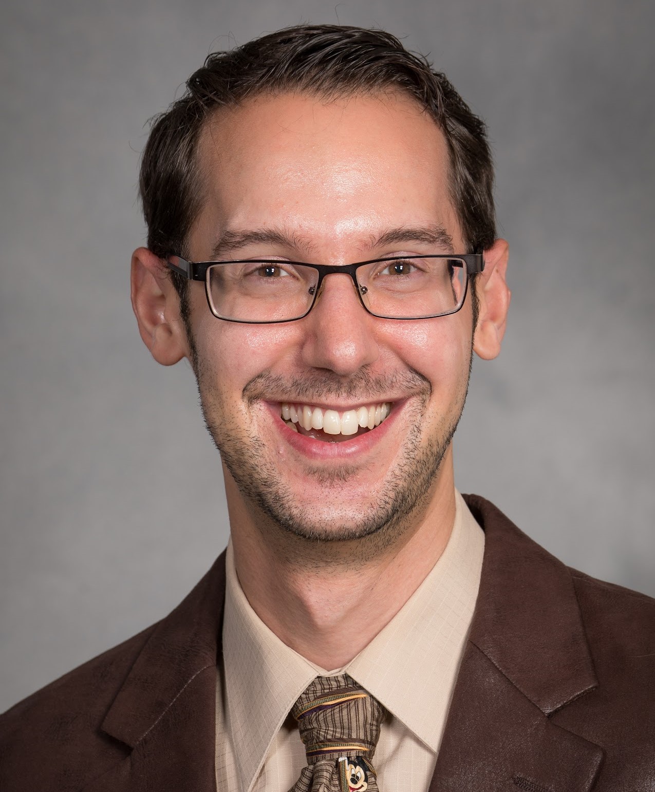 Photo of Dr. Brett Ranon Nachman, smiling man with rectangular glasses wearing a brown suit and patterned tie