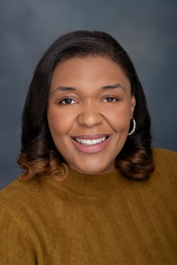 Black woman with curled shoulder length hair wearing a brown sweater smiling at the camera behind a gray background