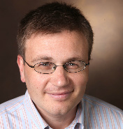 Photo of Zach Warren, white man smiling with rectangular glasses sitting  behind a brown background