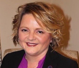 Photo of Jill Rigsby, a white woman with blond curly hair sitting on a beige chair. She wears a black jacket and purple shirt
