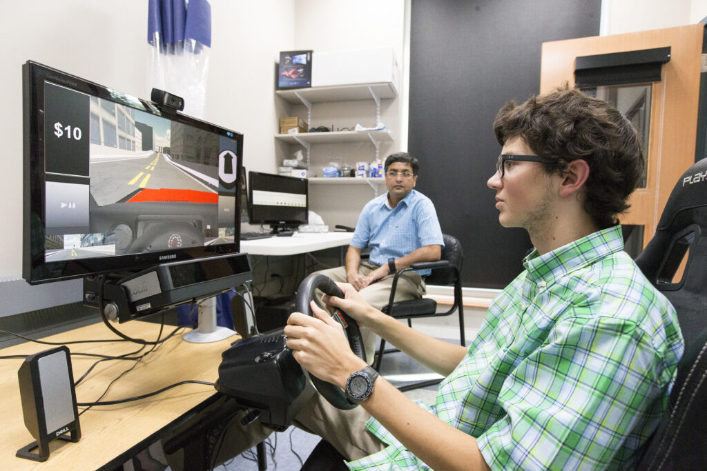 Image of a young man playing a simulation game while a man is looking on from a desk in the background.