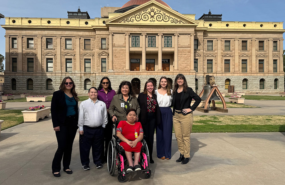 Eight adults stand and sit together outdoors in front of a large historic state capitol building with columns and ornate stonework. The group includes people with and without disabilities; one person sits in a wheelchair at the center. They are dressed in business and business-casual clothing, smiling and posing on a sunny day with the capitol grounds visible behind them