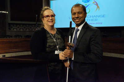 Two people stand indoors in a large room with rows of seats behind them, jointly holding a ceremonial gavel. A projection screen with AUCD branding is visible in the background.