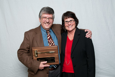 Two people stand together in front of a plain light-colored backdrop. One person holds a wooden award plaque featuring a large ceremonial gavel while the other stands beside them with one arm around their back.