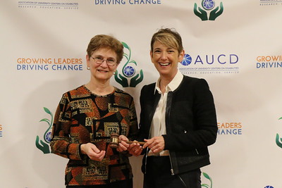 Two people stand in front of a backdrop featuring AUCD logos and the words “GROWING LEADERS DRIVING CHANGE.” They hold a ceremonial gavel together for a photo.