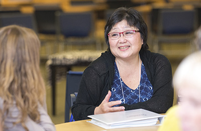 Asian woman with short black hair and glasses sitting at a table talking to another woman.