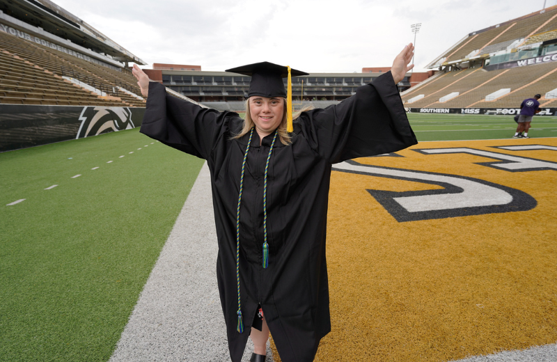 A smiling graduate wearing a black cap and gown stands on a football field with arms raised in celebration. The graduate wears honor cords, and stadium seating and field markings are visible in the background.