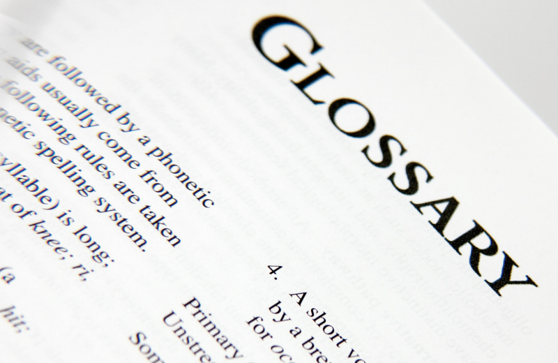 “Close-up, angled photo of a book page with the word ‘GLOSSARY’ in large serif letters at the top, and smaller dictionary-style text visible below on a white page.