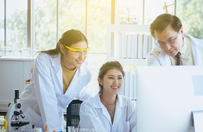 Two happy research students look at a laptop with their professor showing them something on the screen. They are in a lab.