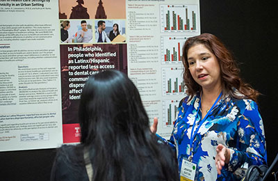 Image of a woman with long brown hair wearing a floral print shirt presenting in front of a poster.