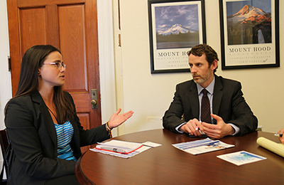 A Latin woman with long brown hair, glasses and suit, speaking to a white man in a suit and tie in a office.