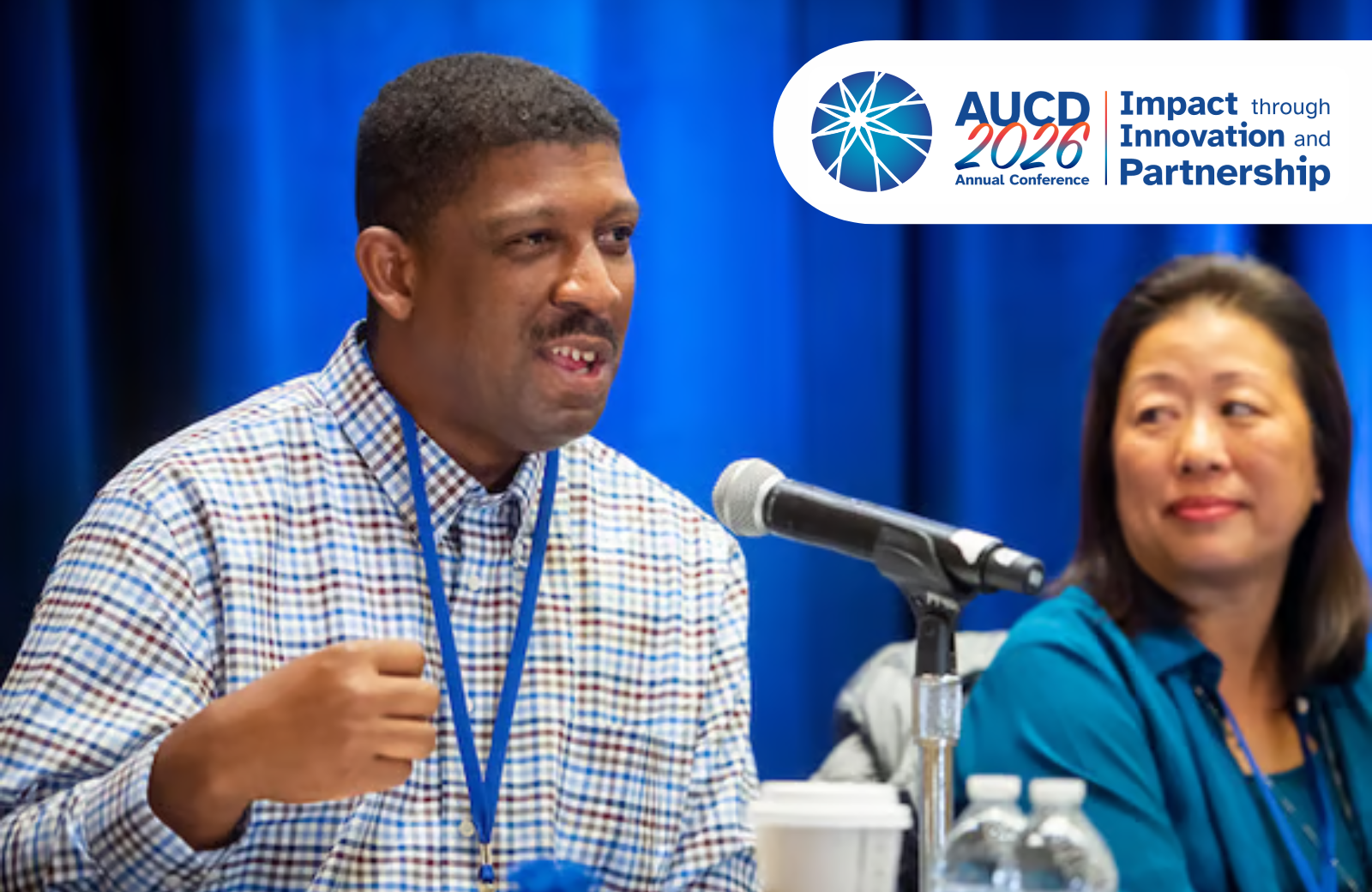 A man in a plaid shirt speaks into a microphone at a conference, with a woman in a teal shirt seated beside him. A banner for the AUCD 2026 Annual Conference is visible in the background.