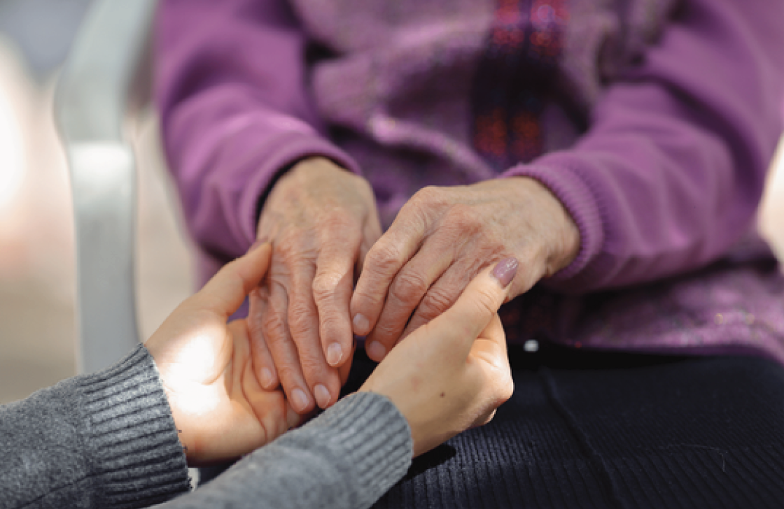A younger person gently holding the hands of an older person, who is wearing a purple sweater and sitting down. 