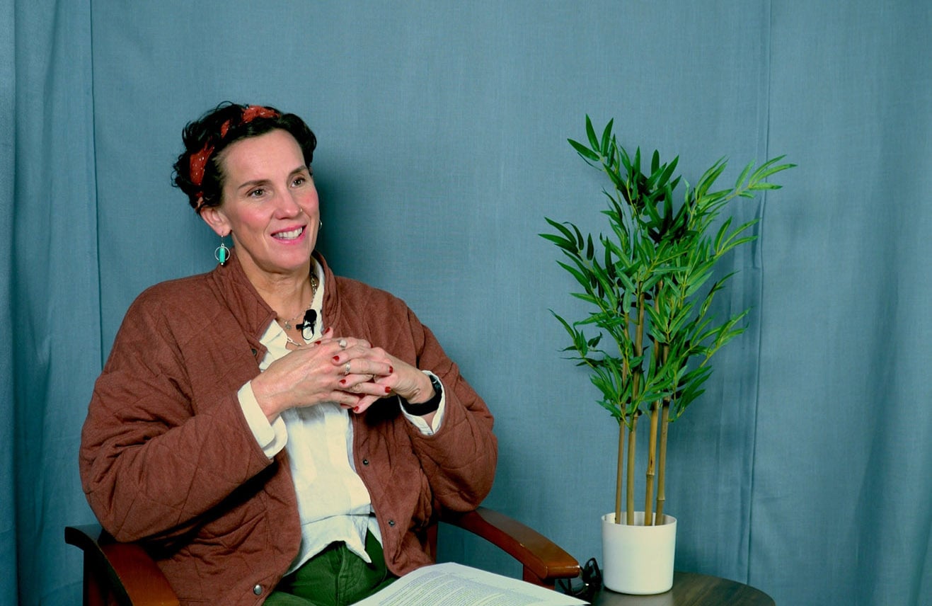 A woman sits in a chair against a blue backdrop, smiling with her hands clasped. She wears a brown quilted jacket, a light shirt, and a patterned headband. A small potted plant sits on a table beside her.