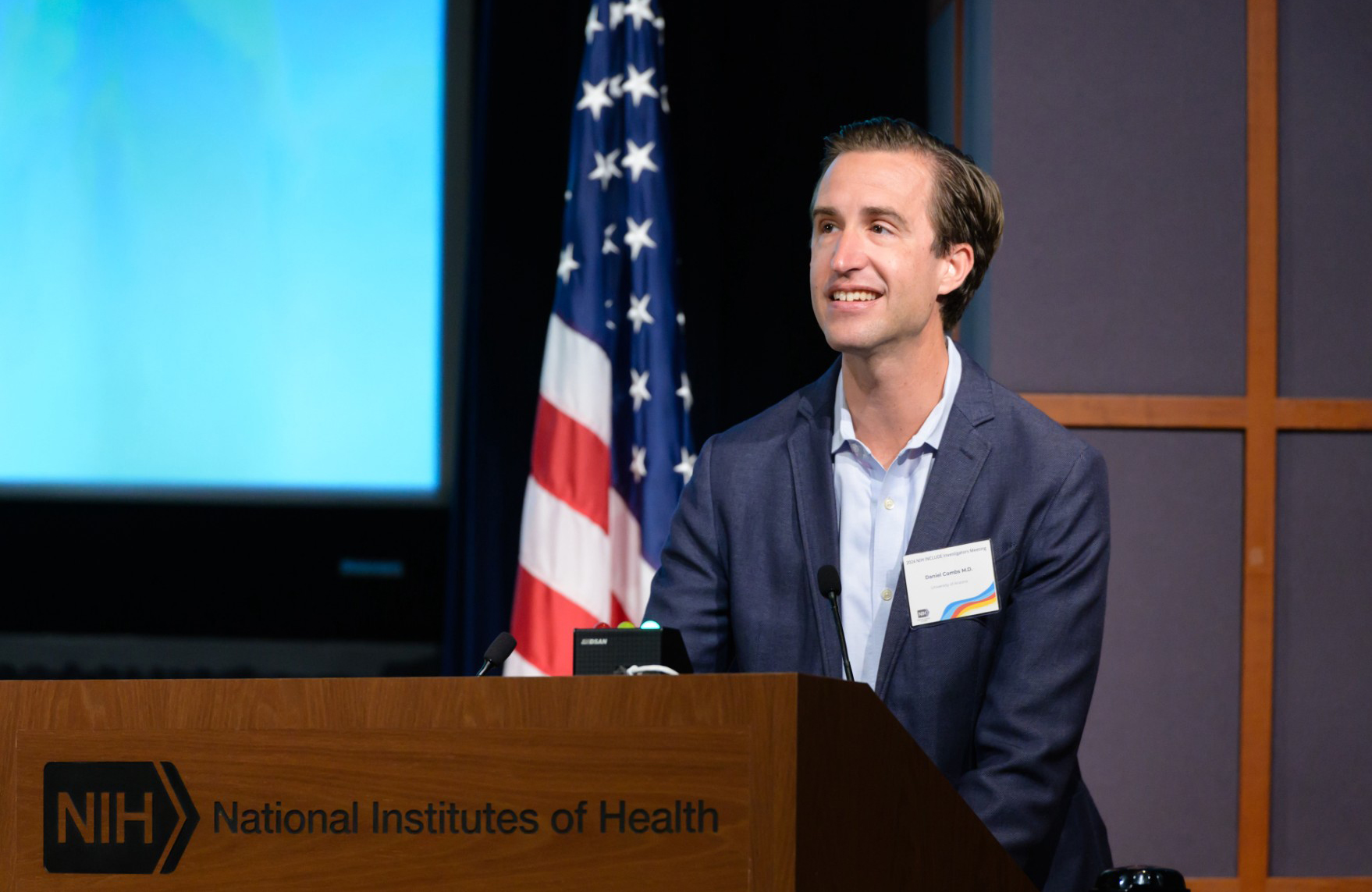 A man speaking at a podium labeled "NIH National Institutes of Health." He is wearing a suit jacket over a light blue shirt and has a name tag.