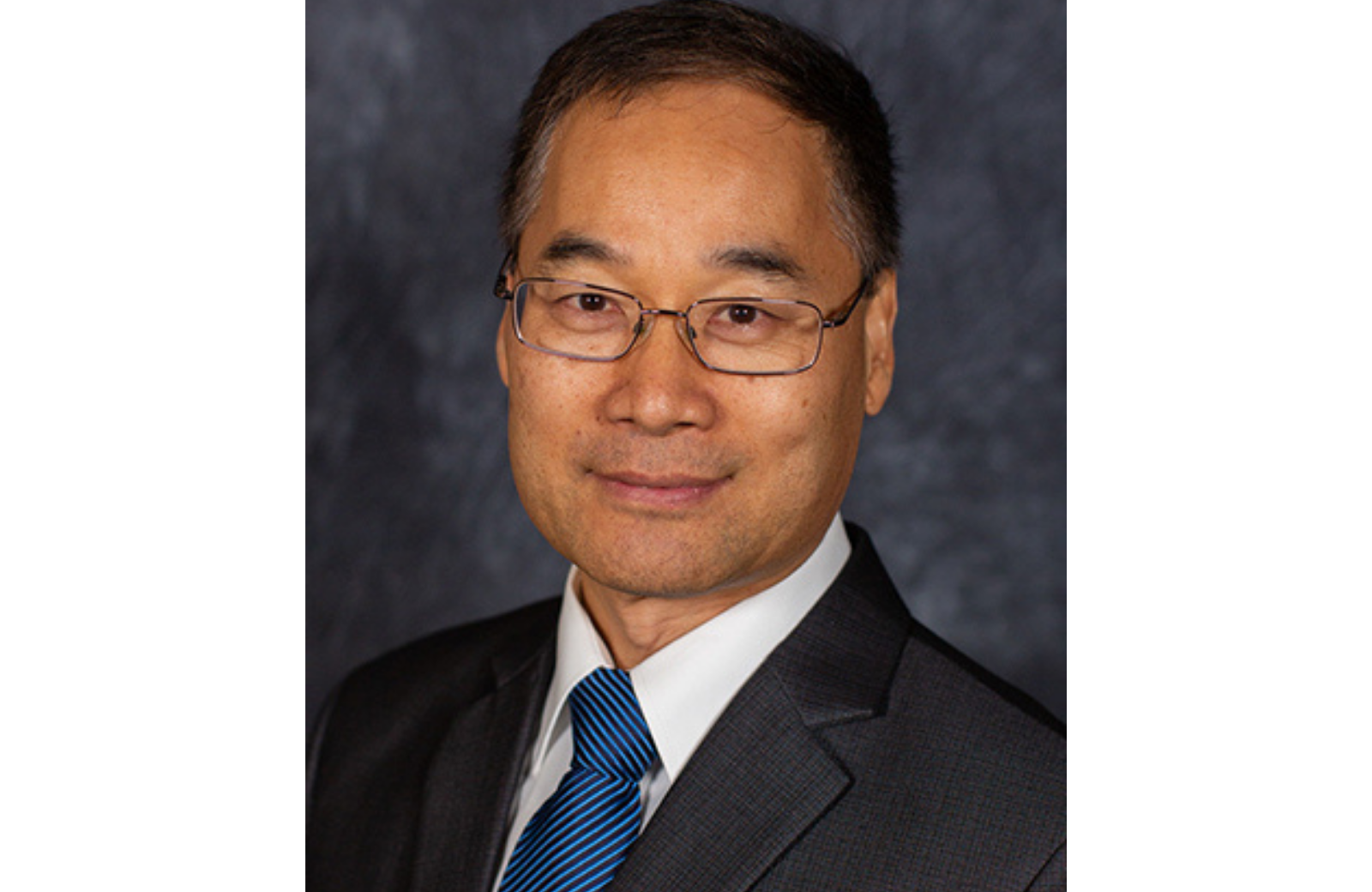 A man wearing glasses, a dark suit, and a blue striped tie smiles at the camera against a dark background.