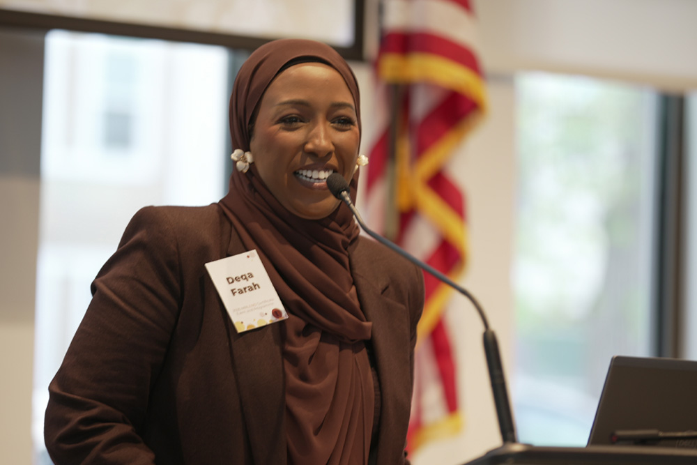 Deqa Farah, a smiling person wearing a brown hijab and matching brown blazer speaks at a podium microphone during an indoor event.