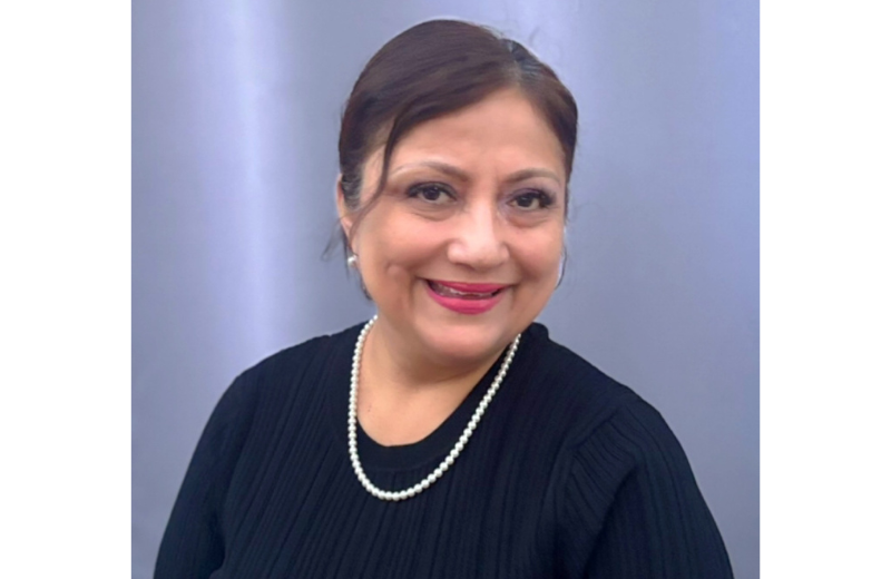 Georgina Georgelos, A person is shown in a portrait-style photograph wearing a long‑sleeved, ribbed black top and a single-strand pearl necklace. The individual has dark hair pulled back and is positioned in front of a smooth gray backdrop.