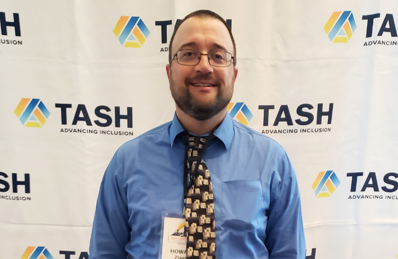  A man wearing glasses, a blue dress shirt, and a patterned tie stands in front of a white backdrop with repeating “TASH – Advancing Inclusion” logos. He is smiling and wearing a name badge that reads “Howard Zlamal.”