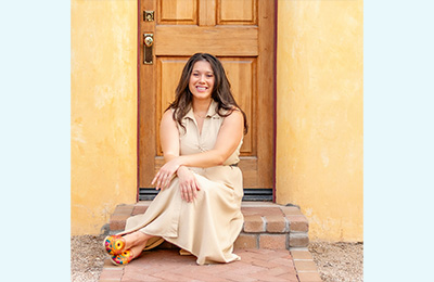Jennifer is pictured in front of a yellow door, wearing a tan dress with long brown hair.