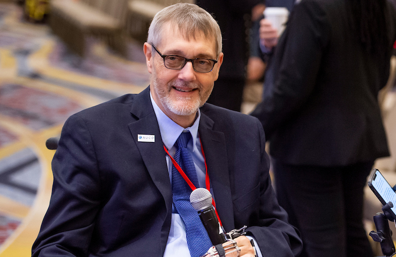 Headshot photo of John Tschida, a smiling man with glasses, graying brown hair and goatee, wearing a navy suit, white shirt, and blue patterned tie, holding a microphone.
