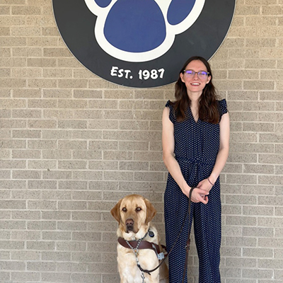 Megan Fayard, a white woman dressed in a jumper with shoulder-length hair and glasses, holding a leash to her guide dog.