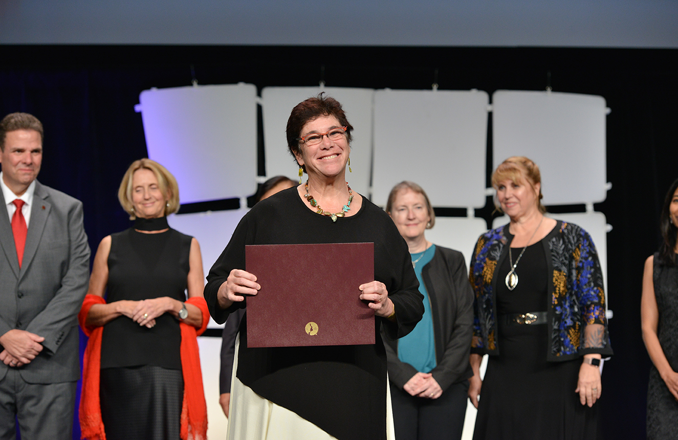 Dr. Fried-Oken stands on a stage holding a brown award folder. She is brightly lit and out in front of a line of professionally dressed people slightly blurred in the background. She has short brown hair, is smiling wide, and is wearing a black and cream dress, pink glasses, and a necklace made of various colored stones.