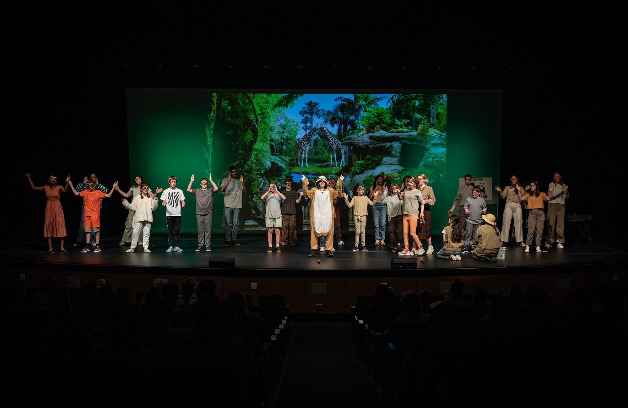 A group of children, teens, and adults stand on a stage in costume, mid-performance of a theatrical production. The backdrop displays a vibrant jungle scene with giraffes, enhancing the immersive setting. The performers are wearing masks and animal-themed attire, with one central performer dressed as a lion. The diverse cast, including individuals with autism, appears to be taking a final bow or celebrating the close of a scene. The audience is visible in the dim foreground, watching attentively.