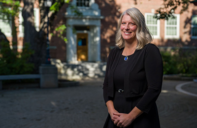 Sarah Smith is a woman with wavy blonde hair, wearing a nice black dress and cardigan. She's posing in front of a brick building on the UNH campus.