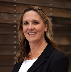 Sheli Reynolds, a white woman with shoulder length hair, wearing a suit and smiling at the camera.