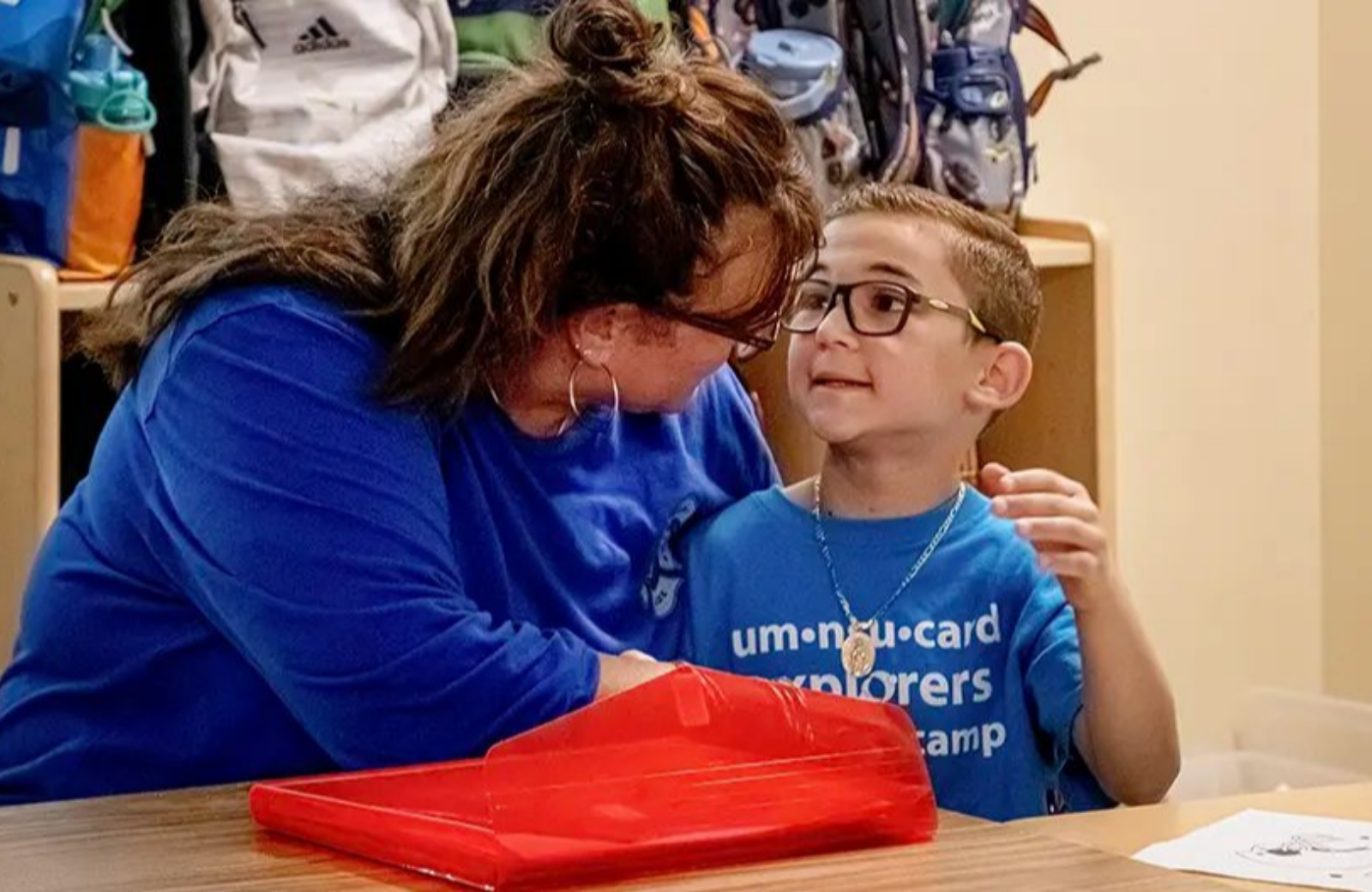 A young child wearing glasses and a blue camp T-shirt sits closely with an adult in a classroom, making warm eye contact as they talk. The adult, also wearing a blue shirt and glasses, leans in attentively with one arm around the child in a supportive, reassuring posture. A bright red plastic tray rests on the table in the foreground, with a sheet of paper nearby.