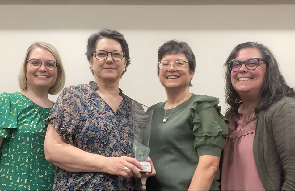 Four women standing together, smiling at the camera. The second person from the left is holding a clear glass or acrylic trophy.