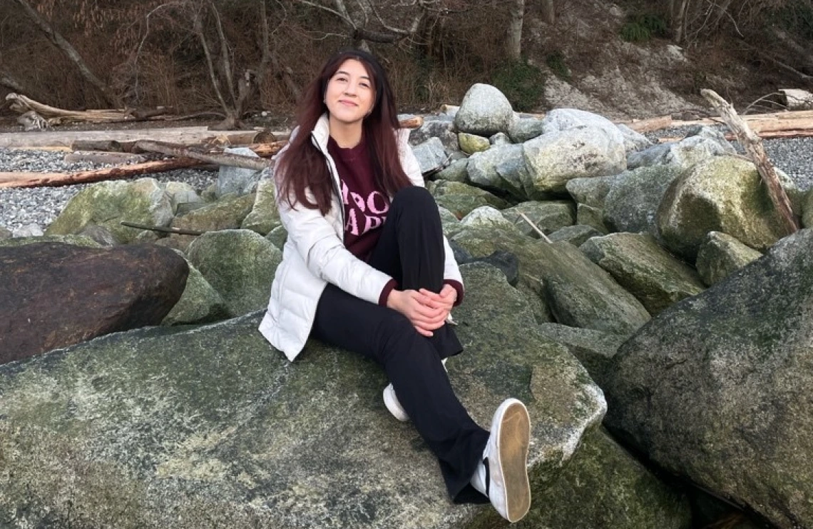 Anahy Fernandez sitting on a large rock smiling. Anahy has long red-brown hair, wearing winter wear, and is holding on to one leg while the other is extended.