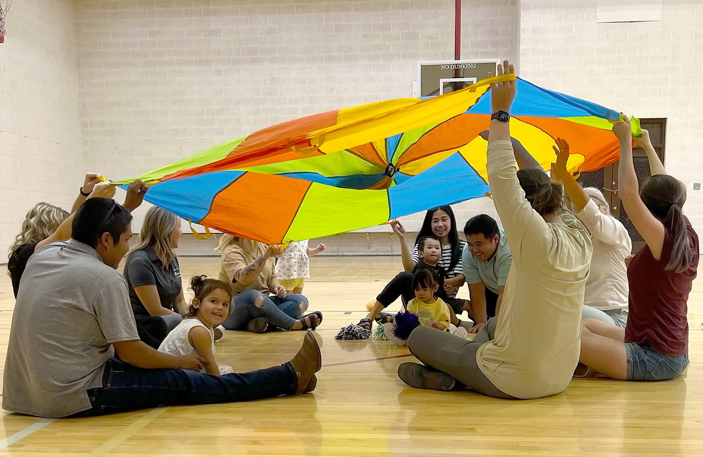 Children siting in a circle with adults that are holding a parachute over their heads 