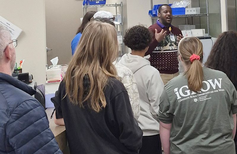 A small group of people stands in a clinic or lab space, facing a speaker who is talking and gesturing with his hands. Shelving with medical or lab supplies is visible in the background. One person in the group is wearing a gray shirt with “iGROW – Generating Rural Opportunities for Work” printed on the back. The group appears to be listening attentively during a tour or presentation.