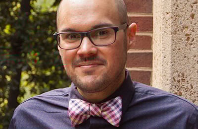 Headshot of Pablo Juárez, a man with shaved head and goatee, black framed glasses, an indigo button-down shirt, and a pink plaid bow tie.