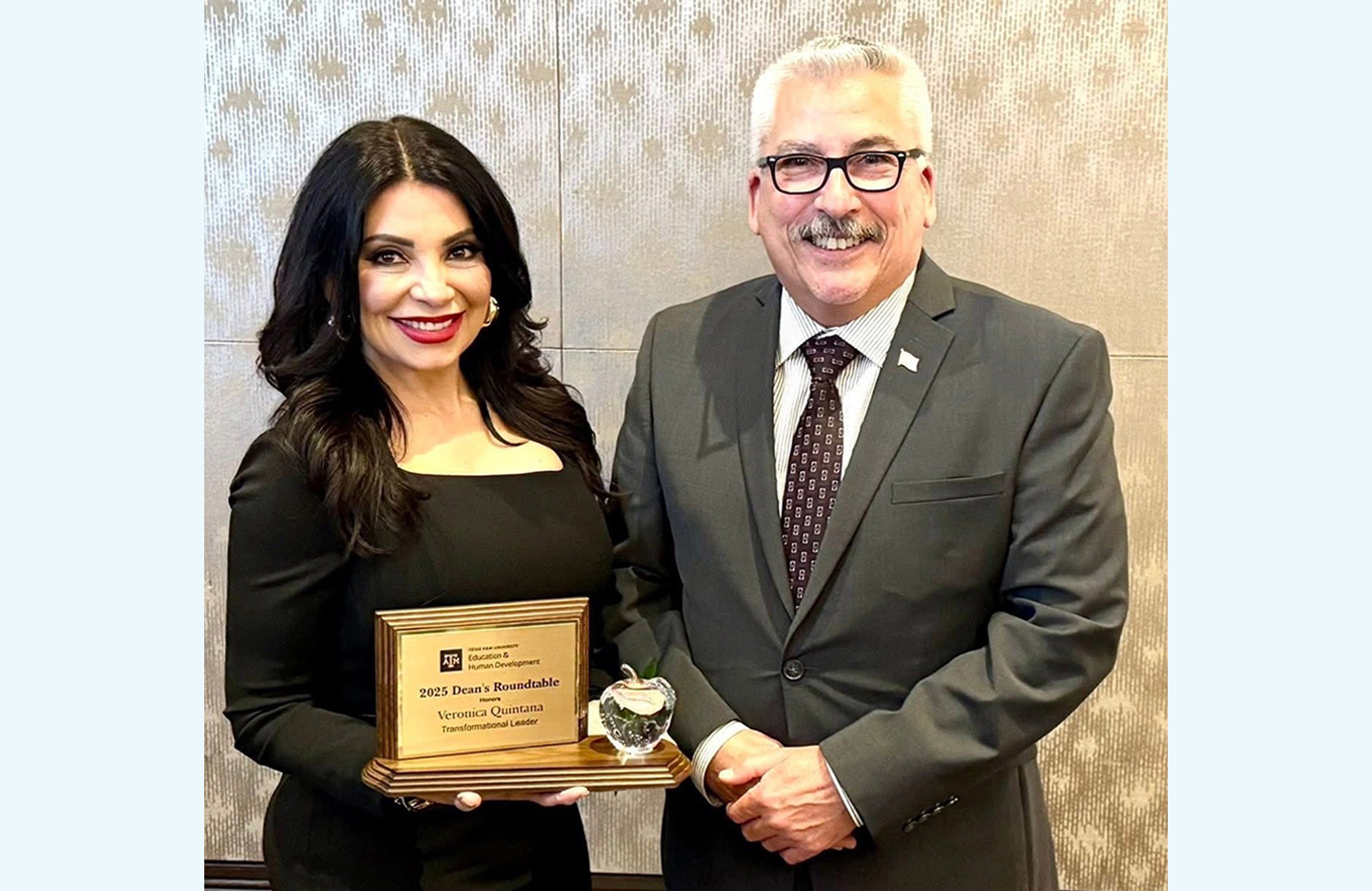 The image shows two individuals standing side by side, smiling at the camera. The woman on the left has long, dark wavy hair and is dressed in a black dress. She is holding a wooden plaque that reads: Texas A&M University College of Education & Human Development 2025 Dean’s Roundtable honors Veronica Quintana Transformational Leader There is also a small glass apple trophy on the plaque base. The man on the right is wearing glasses, a grey suit, a patterned tie, and an American flag pin on his lapel. They are standing in front of a neutral, textured wall.