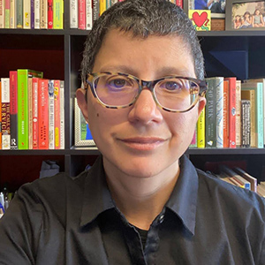 Image of Anna Costalas, a smiling olive skinned queer with short greyish black hair and brown eyes, wearing tortoise glasses and a button down shirt with a book filled bookcase in the background.