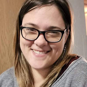 Image of Melanie Davis, a smiling white woman with long brown hair and glasses wearing a grey sweater.