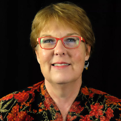 Image of Denise M. Rozell, a white woman with short redish blonde hair, she is wearing red glasses and a floral suit. 