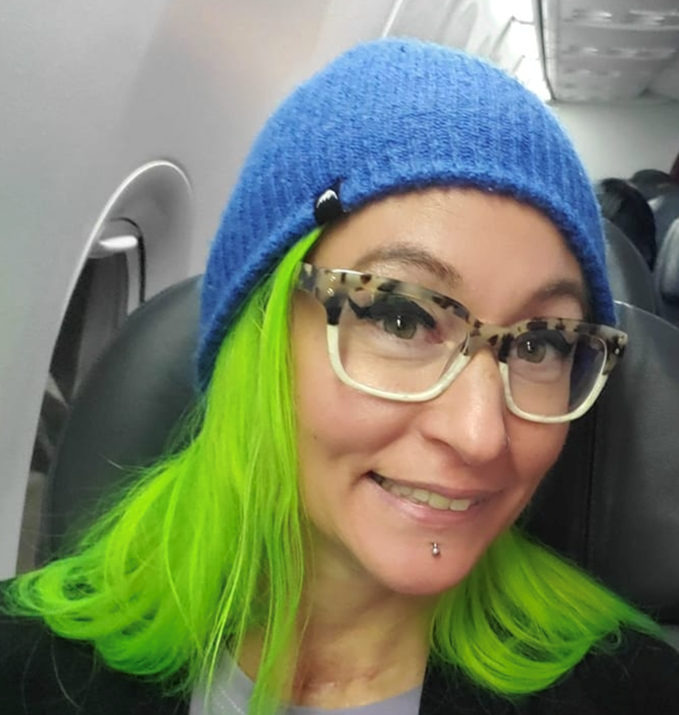 woman with bright green hair and brown and tan dotted glasses wearing a blue beanie hat smiling